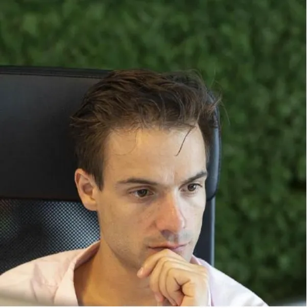 A man with short brown hair sits in a chair, resting his chin on his hand, appearing deep in thought, with a green leafy background.