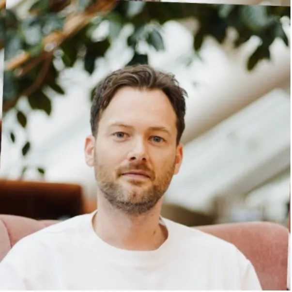 A person with short hair and a beard, wearing a white shirt, seated indoors with plants and soft lighting in the background.