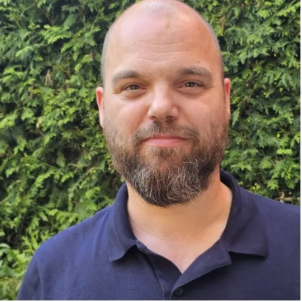 A man with a beard and balding head wearing a navy shirt stands in front of green leafy bushes, smiling slightly at the camera.