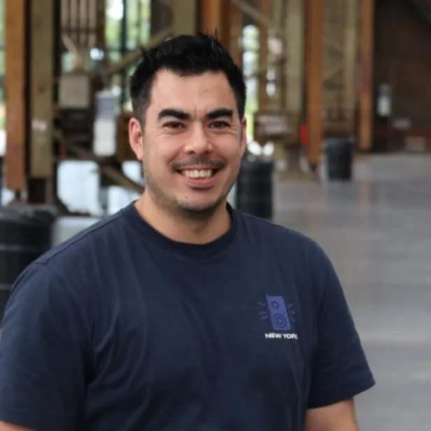 A man in a navy T-shirt with a speaker design smiles in an industrial-style setting with wooden beams and a concrete floor.