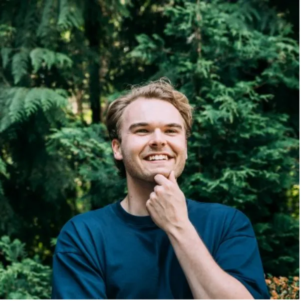 A smiling man in a blue shirt poses thoughtfully with his hand on his chin, surrounded by lush green foliage.